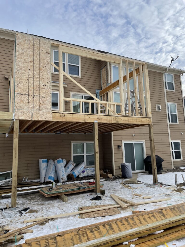 A new wooden two-story addition frame attached to the back of a tan house, featuring exposed wall studs and OSB sheathing.