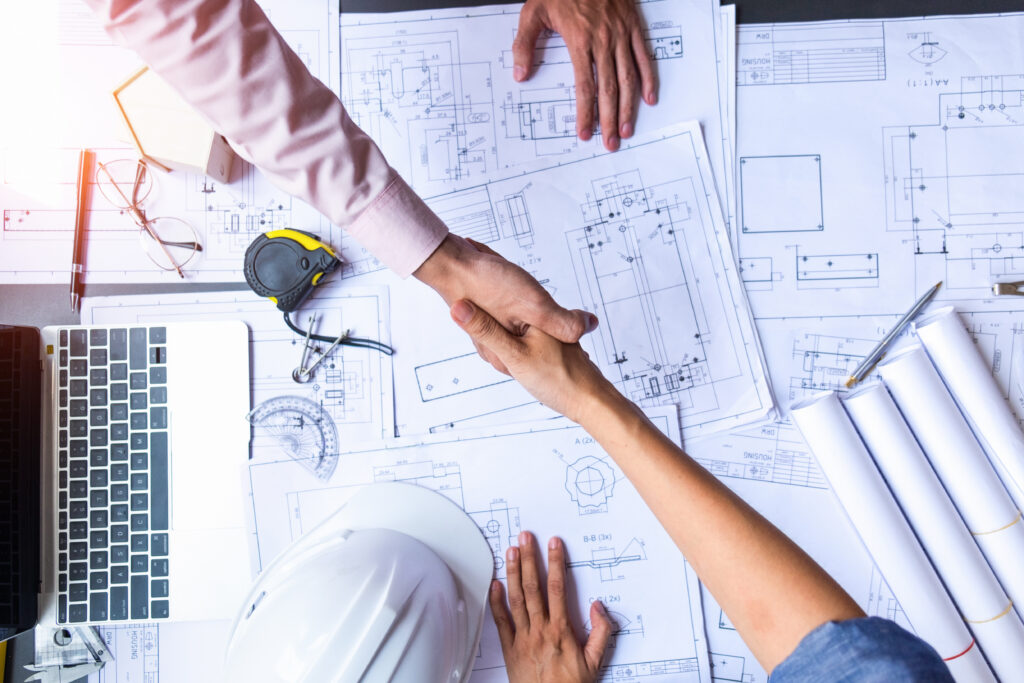 Two people shaking hands over architectural blueprints on a table.