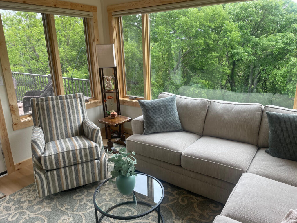 Modern living room with beige sectional sofa, striped armchair, glass coffee table, and large windows overlooking a lush green forest.